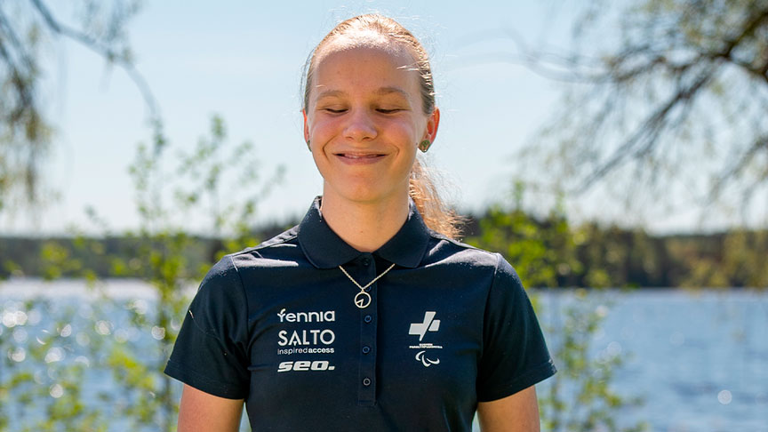 Ronja Hampf hymyilemässä kameralle. Taustalla vihreää kasvustoa ja vesistö. Aurinkoinen sää, ulkona.